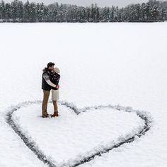 a man and woman standing in the middle of a heart shaped snow covered field with their arms around each other