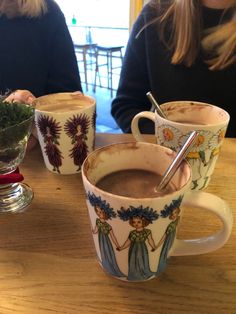 two women sitting at a table with cups of coffee in front of them, one holding a spoon