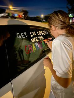 a woman writing on the side of a car that says get in losers we're graduate