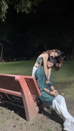 two people sitting on top of a bench in the park at night with trees and grass behind them