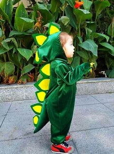 a little boy in a green dinosaur costume standing on the sidewalk with his hand up to his face