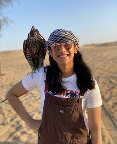 a woman holding an owl on her arm in the desert with sand and trees behind her