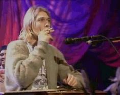 a man standing in front of a microphone on top of a wooden table next to a purple curtain