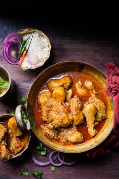 two bowls filled with chicken curry next to rice and red onions on a wooden table