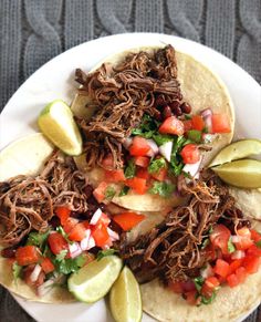 three shredded beef tacos on a white plate with lime wedges and sliced avocado