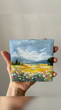 a hand holding up a small piece of art with flowers in the foreground and clouds in the background