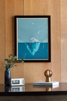 Waverider gift lamp sits atop a book on a metal desk. A modern flip clock and vase with botanicals are next to it. Behind the desk is a wood paneled wall with a framed piece of art.