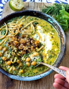 someone is holding a spoon over a bowl of soup with noodles and greens on the side