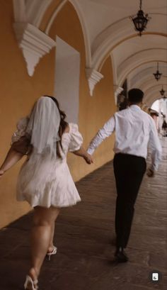 a man and woman dressed in white walking down a hall way with one holding the other's hand