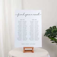 a seating chart on top of a table next to a potted plant and white curtain