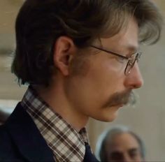 a man with glasses and a moustache looks off into the distance while standing in front of other people