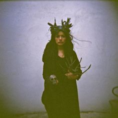 a woman dressed in black holding scissors and wearing a horned headdress, standing against a white wall