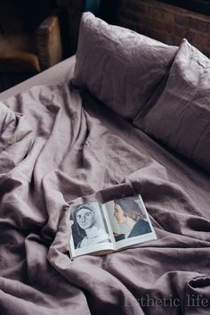 an open book sitting on top of a bed next to pillows and blankets in front of a brick wall