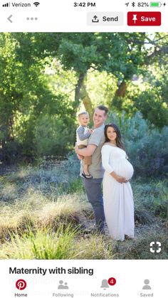 a man and woman holding a baby while standing in the grass with trees behind them