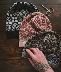 a person's hand next to three knitted hats on a wooden table with scissors