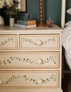 a white dresser with green leaves painted on the drawers and flowers in vases next to it