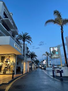 an empty street with palm trees and shops on both sides at dusk in front of the ocean