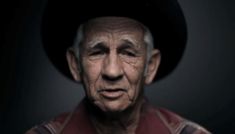 an old man wearing a black hat and red shirt is looking at the camera while standing in front of a dark background