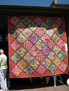 two people standing next to a large quilt on the side of a building with one person looking at it