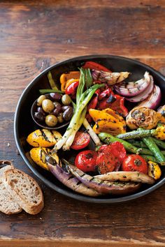 a pan filled with different types of vegetables on top of a wooden table next to bread