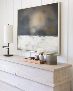 a large painting hangs on the wall above a dresser in a white room with drawers