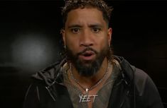 a man with long hair and piercings on his chest wearing a black jacket, standing in front of a dark background