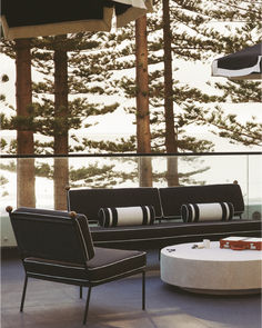 A chic outdoor lounge setup featuring a sleek black sofa and matching chair with elegant white piping and round pillows, set against a backdrop of tall trees and a serene coastal view. Outdoor Comfort, Elegant Sofa, Black Sofa, Hammock Chair