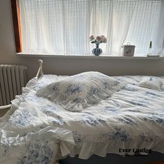 an unmade bed in front of a window with white curtains and blue flowers on it