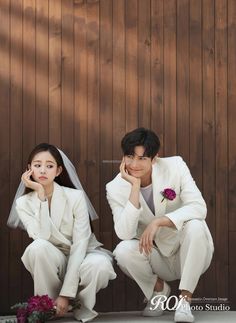 a man and woman dressed in white sitting on the ground next to a wooden wall