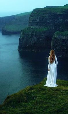 a woman standing on top of a lush green hillside next to the ocean with cliffs in the background