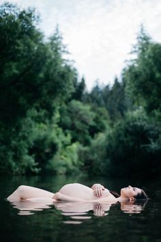 a naked woman floating on top of a body of water next to lush green trees