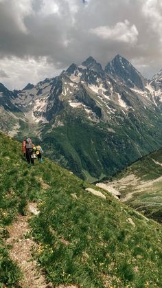 two people hiking up the side of a mountain with backpacks on their back, in front of some snow capped mountains