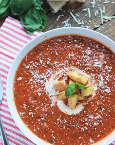 a bowl of tomato soup with bread and basil on the side, ready to be eaten