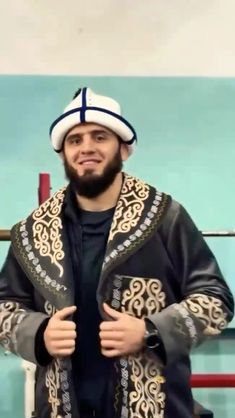 a man with a beard wearing a black jacket and white hat standing in front of a blue wall