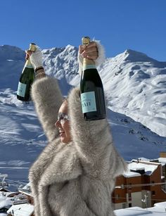 a woman holding two bottles of wine in the air while standing on top of a snow covered mountain