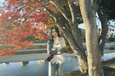 a woman standing next to a tree in front of a lake with red leaves on it