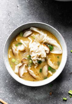 a white bowl filled with dumplings next to green onions and chopsticks on a gray surface