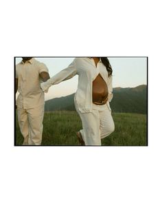 two people in white suits are walking through the grass with mountains in the background and one person is holding her hand out