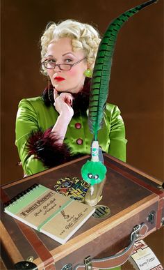 a woman sitting at a table with a green feather on it's head and an old suitcase in front of her