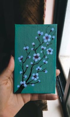 a hand holding up a small painting with blue flowers on green canvases in front of a window