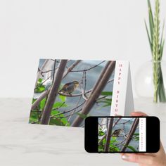 a person holding up a card with a bird on it next to a vase filled with flowers