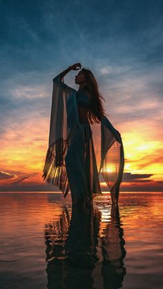 a woman standing in the water at sunset with her arms around her neck and wearing a sheered shawl