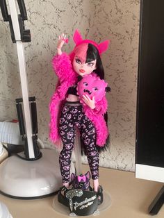 a doll with pink hair and black pants holding a stuffed animal in front of a computer monitor