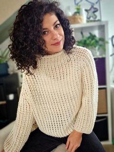 a woman with curly hair sitting on a couch wearing a white sweater and black pants