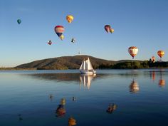 many hot air balloons are flying over the water and hills in the distance, while a sailboat is floating on the lake