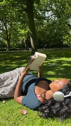 a woman laying in the grass reading a book and listening to headphones on her head