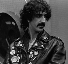 a man wearing a leather jacket and moustache with patches on his chest, standing in front of an airplane