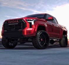 a red truck parked on top of a parking lot next to the ocean at sunset