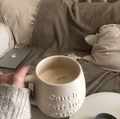 a person holding a coffee mug in front of a laptop computer on a couch with pillows