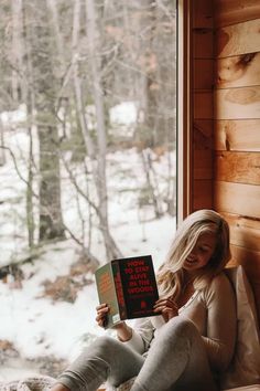 a woman sitting on a window sill reading a book in front of a snowy forest